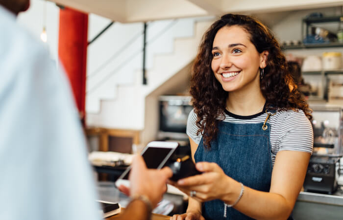 cashier with curly brown hair wearing a denim apron helps a customer from across the counter with paying for their purchase with their phone